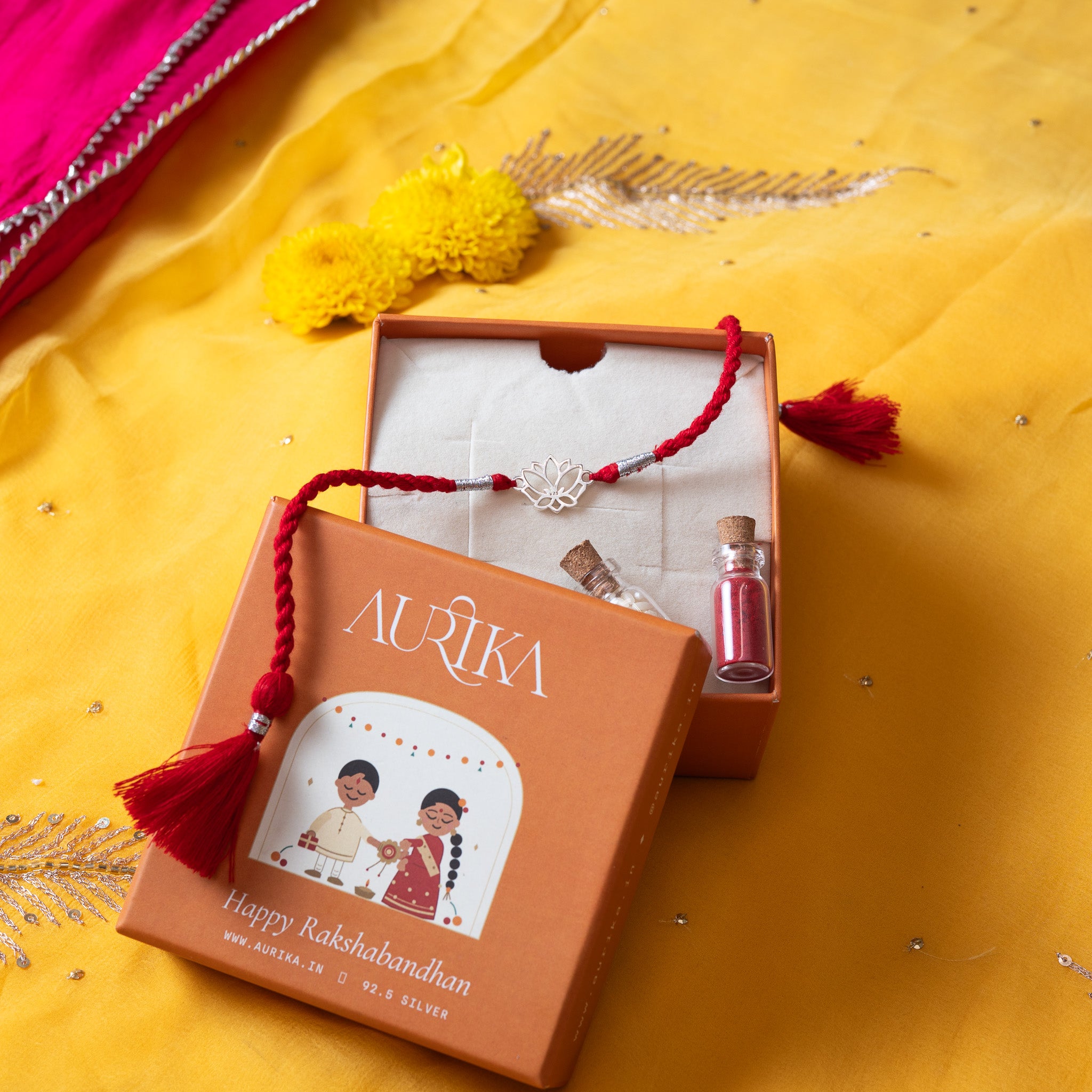 Lotus rakhi with its red cord in an open Aurika gift box with roli and akshat vials