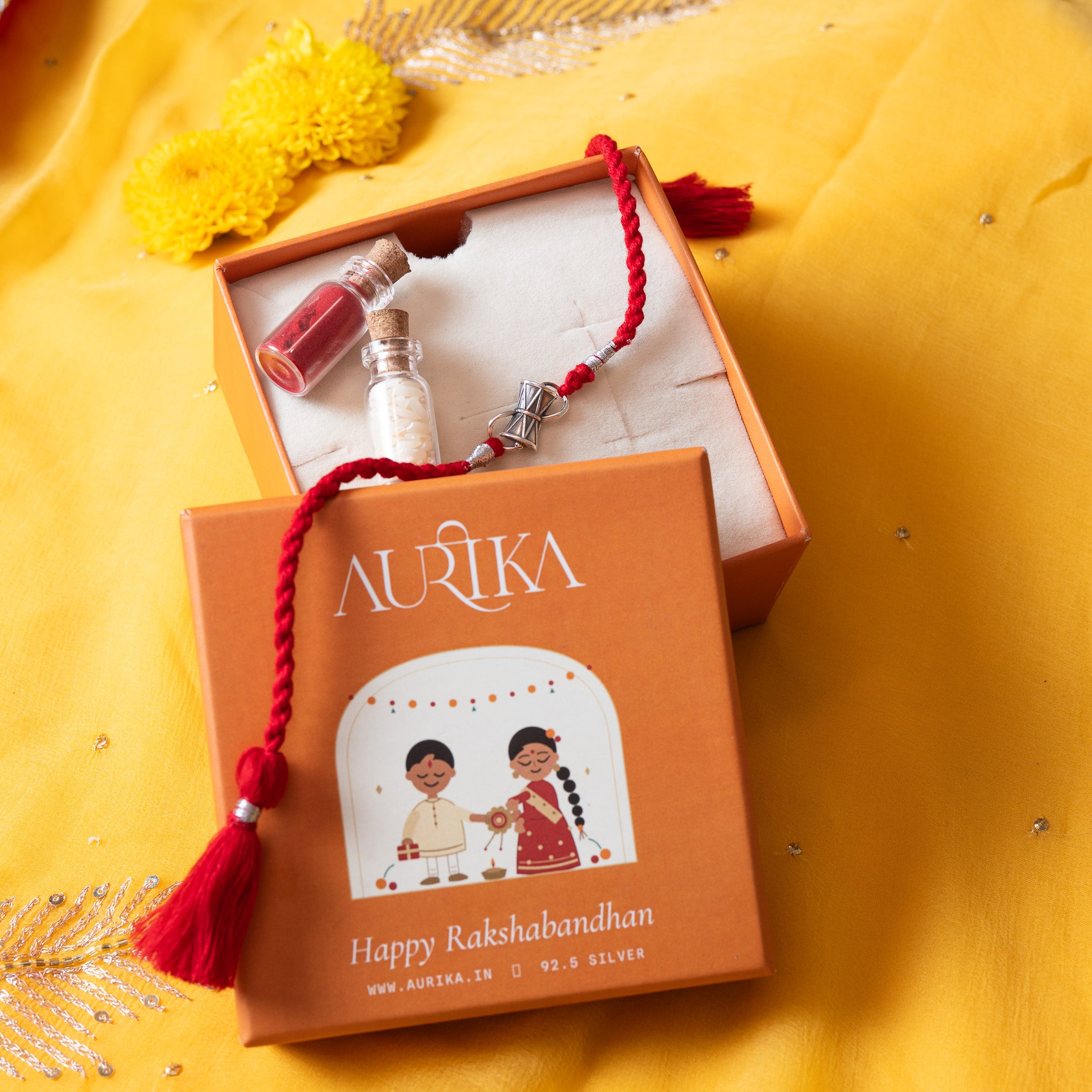 Damaru drum rakhi with its red cord in an open Aurika gift box with roli and akshat vials