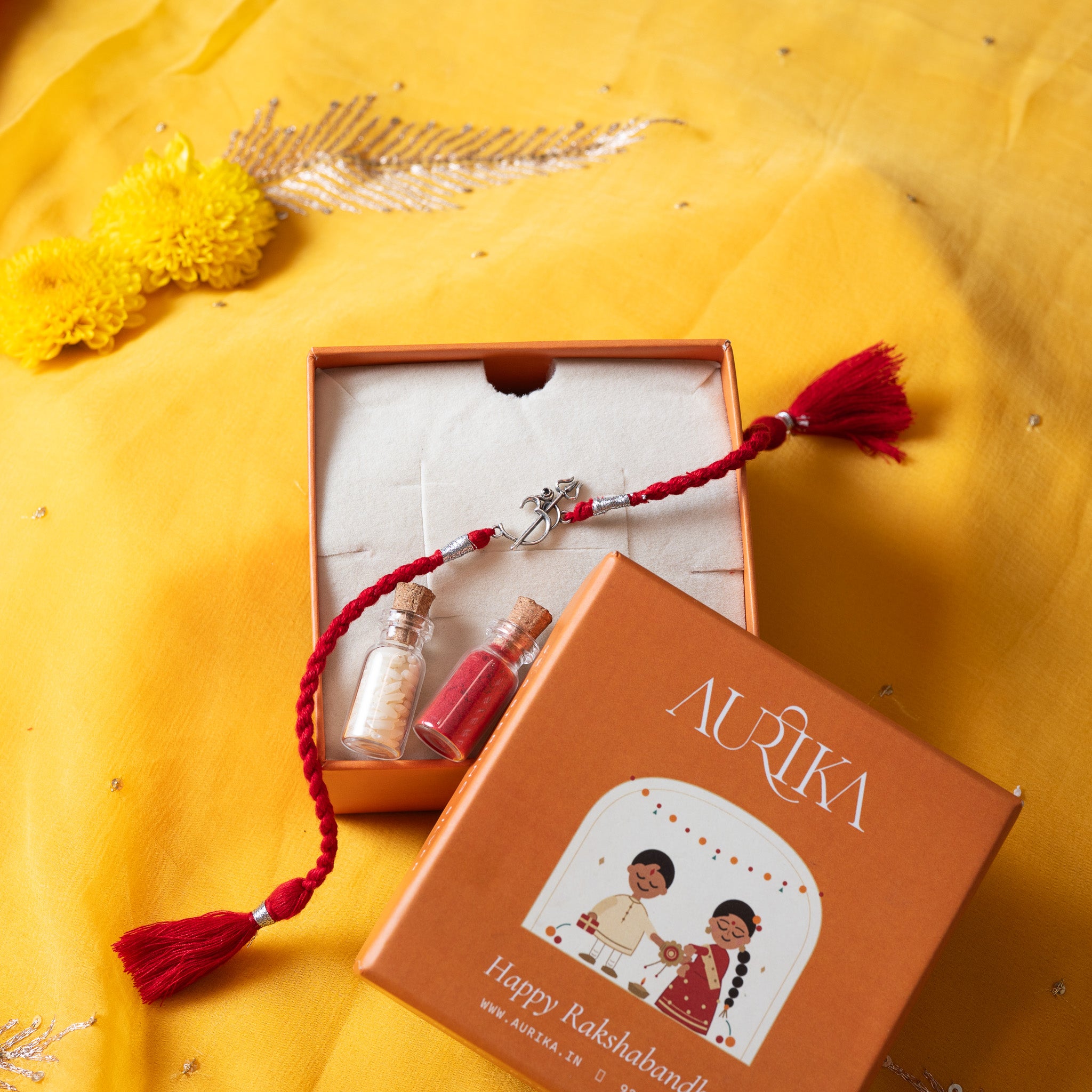 Om trishul rakhi with its red cord in an open Aurika gift box with roli and akshat vials