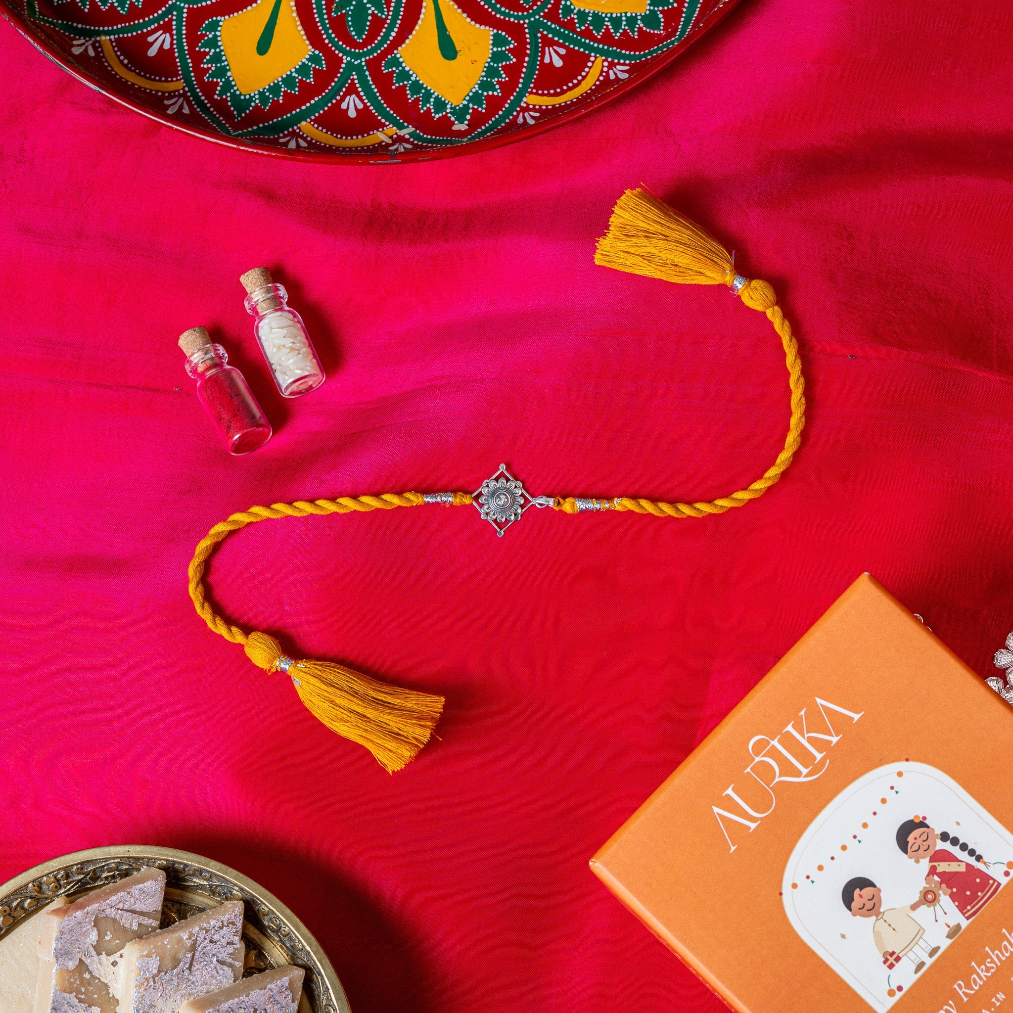 Champak flower rakhi in 925 silver on a marigold yellow silk cord, on rani pink cloth