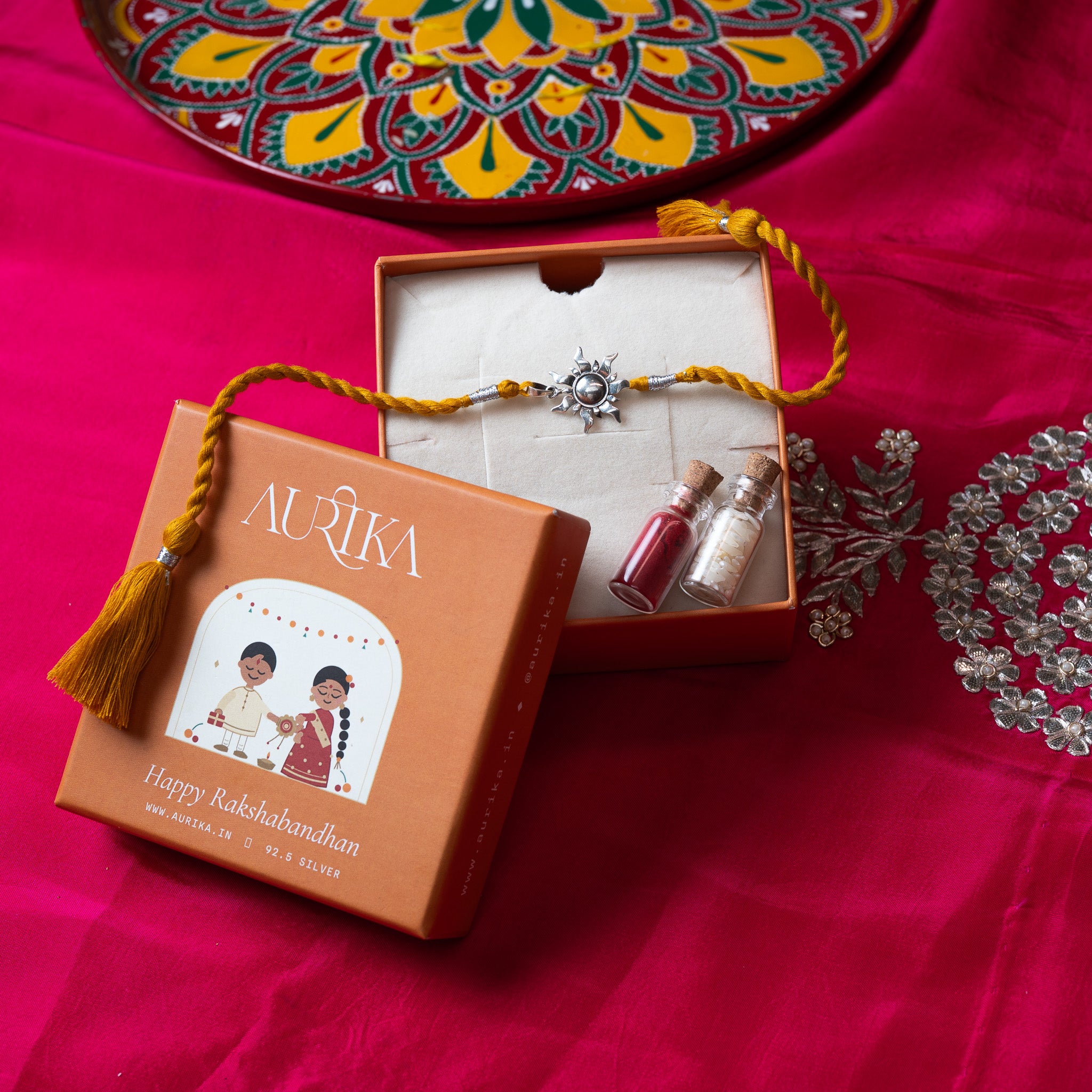 Surya sunburst rakhi with its marigold cord in an open Aurika gift box on rani pink cloth