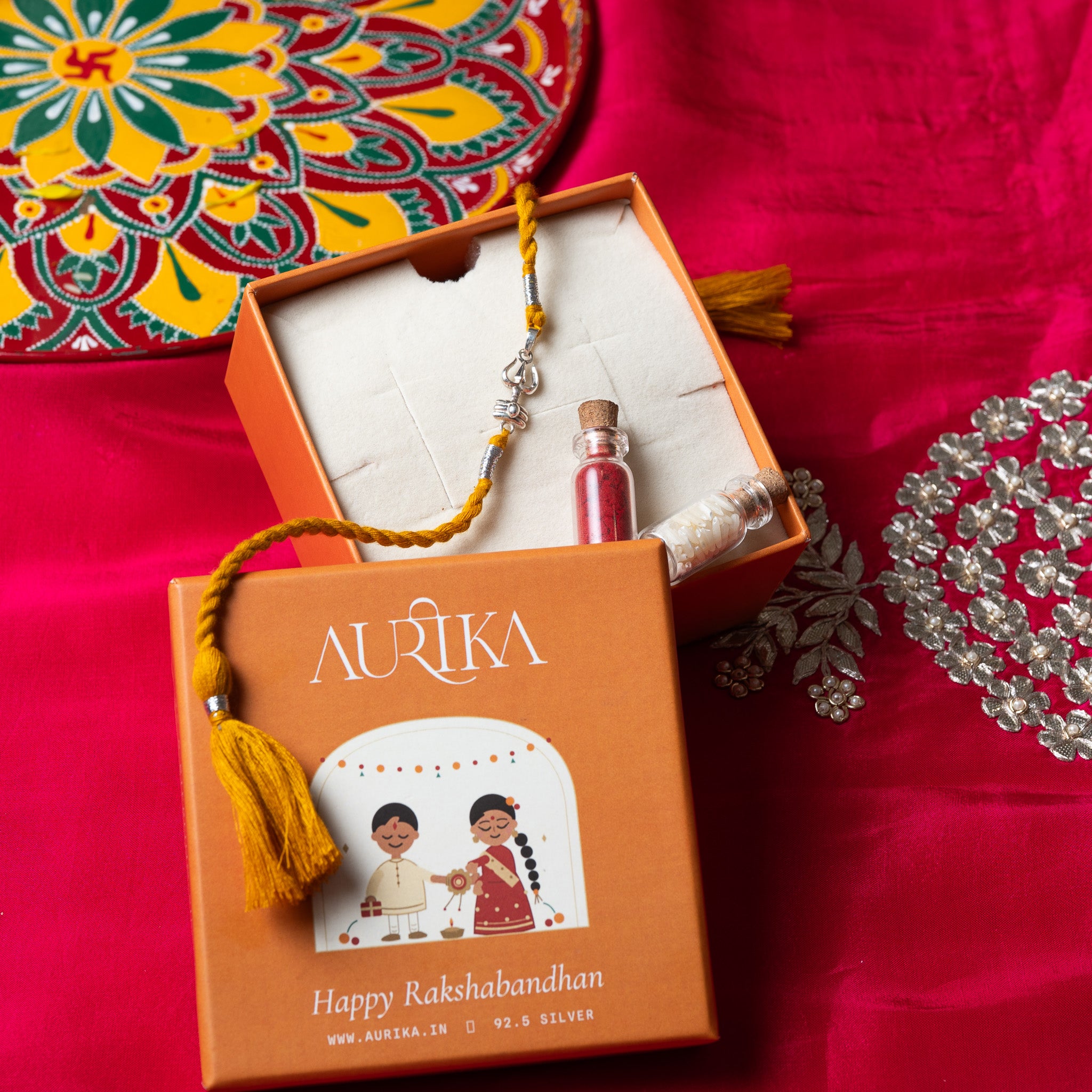 Trishul and shivling rakhi with its marigold cord in an open Aurika gift box on rani pink cloth