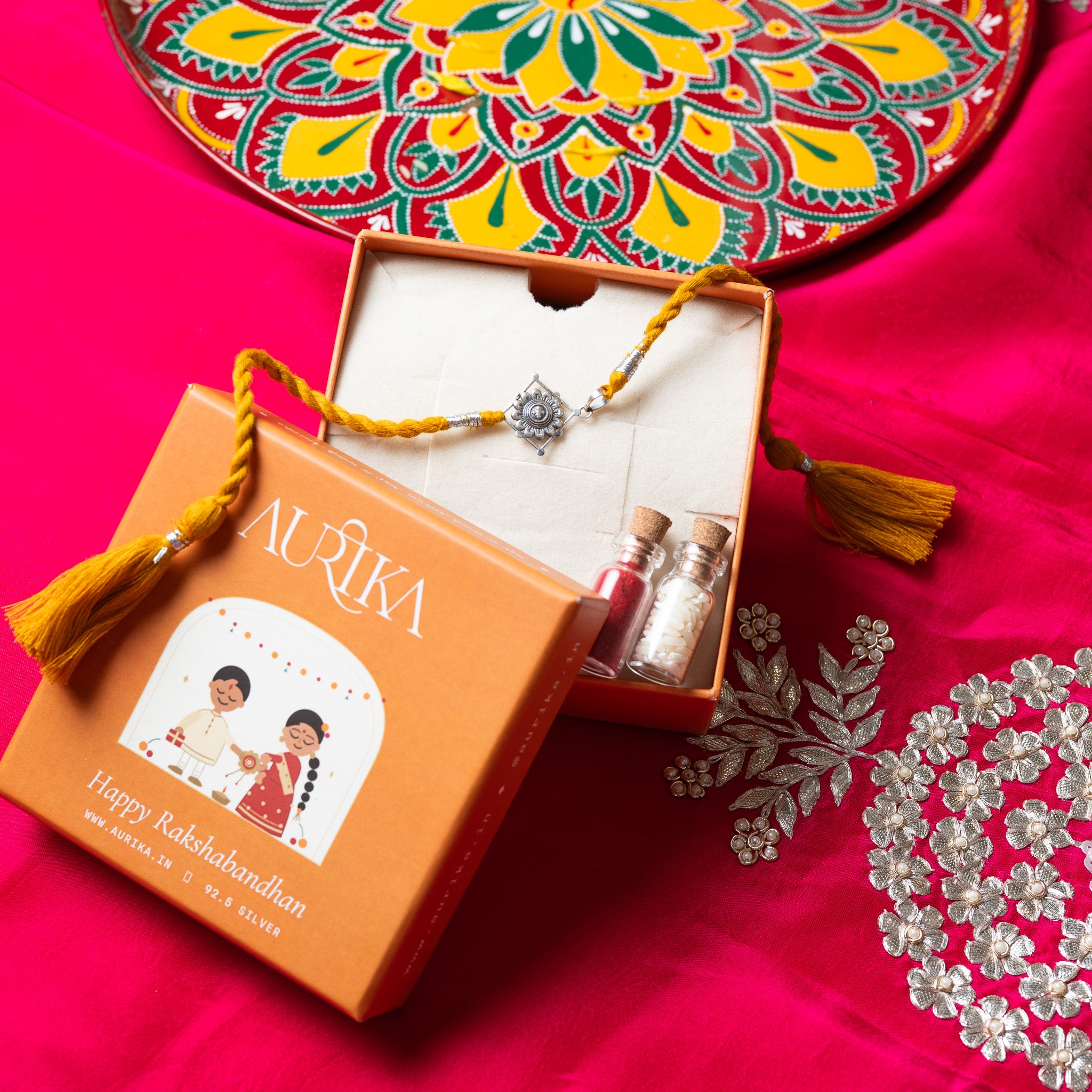 Champak flower rakhi with its red cord in an open Aurika gift box with roli and akshat vials