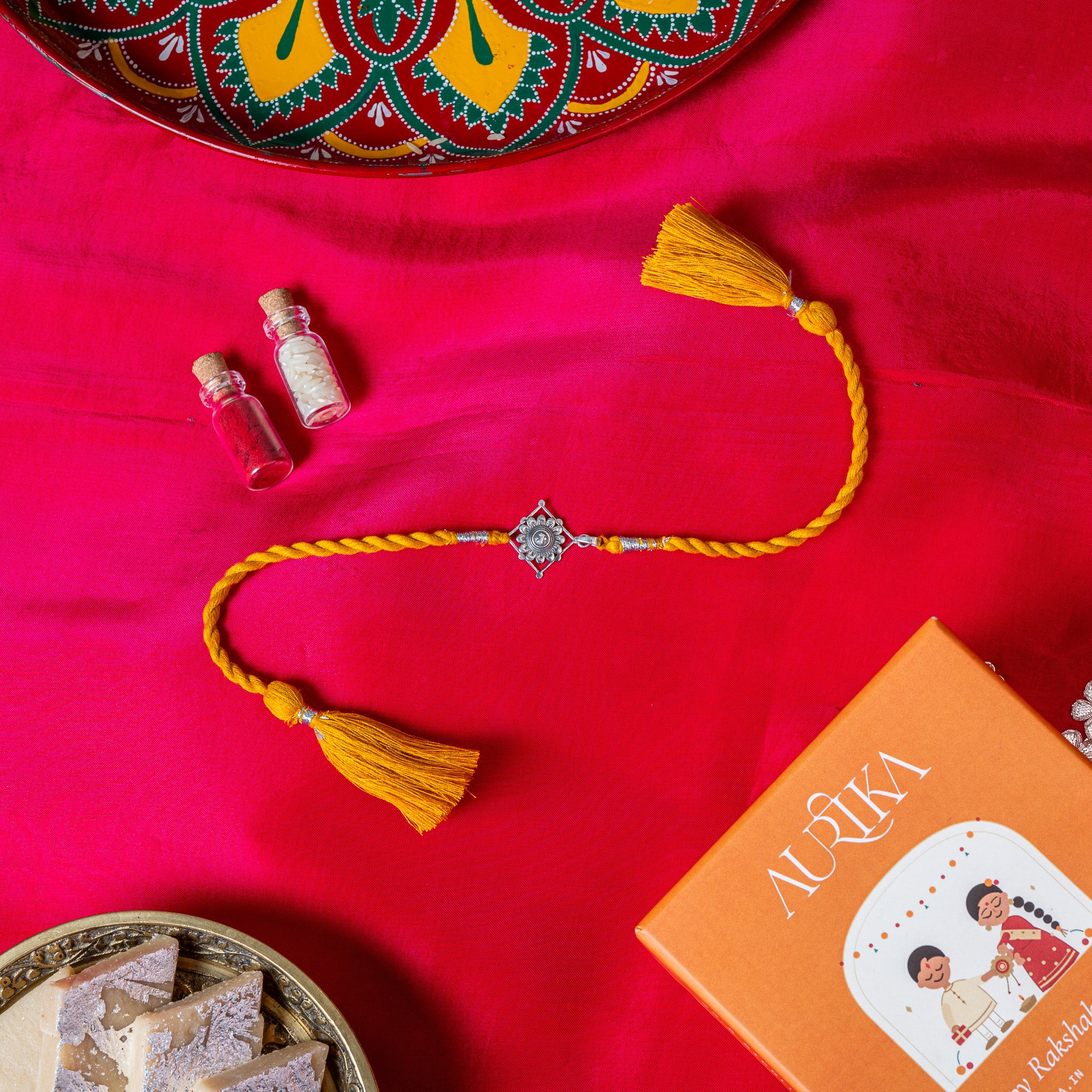 Champak flower rakhi in 925 silver on a marigold yellow silk cord, on rani pink cloth