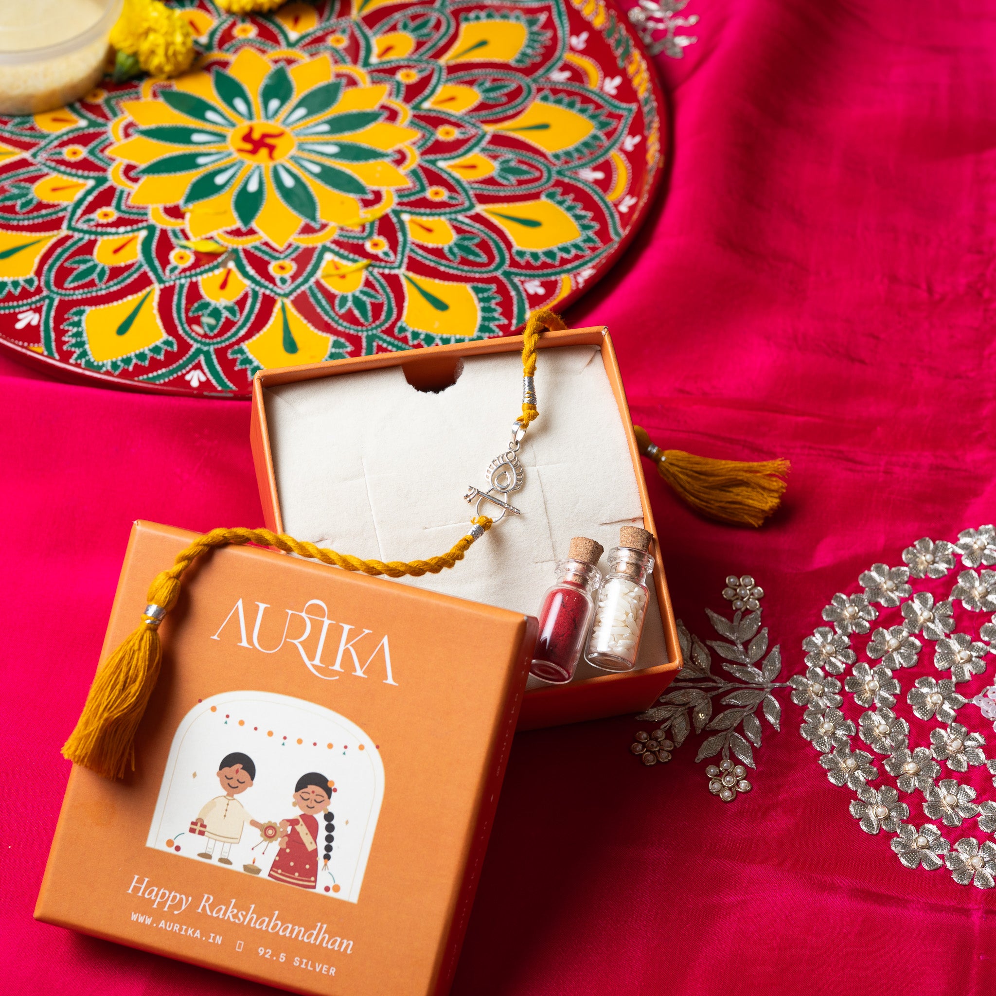 Bansuri and peacock feather rakhi with its red cord in an open Aurika gift box with roli and akshat vials