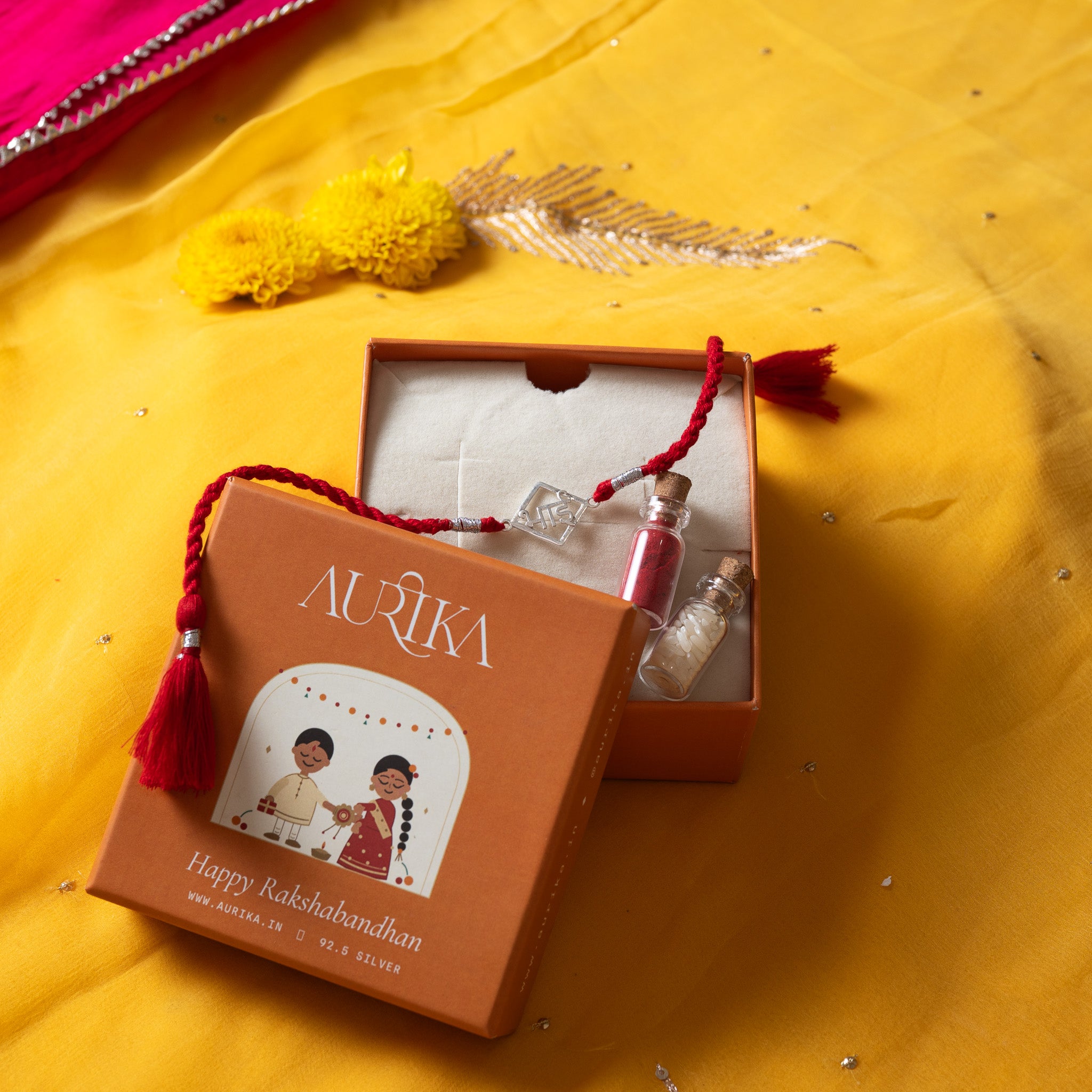 Bhai lettering rakhi with its red cord in an open Aurika gift box with roli and akshat vials
