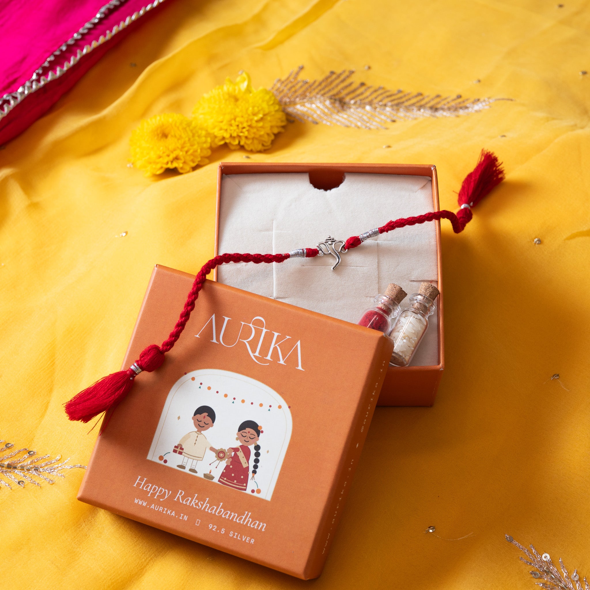 Ganesha rakhi with its red cord in an open Aurika gift box with roli and akshat vials