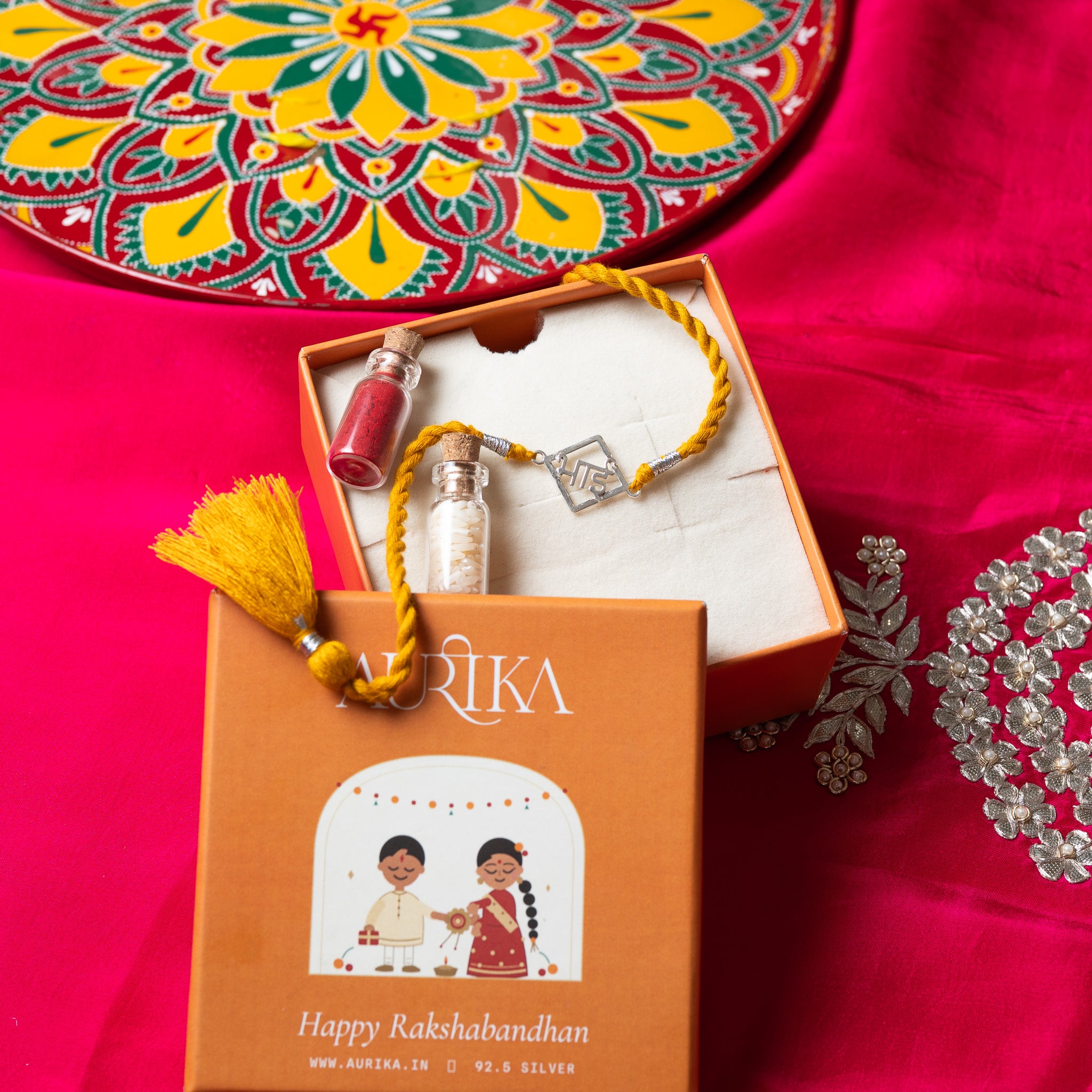 Bhai lettering rakhi with its marigold cord in an open Aurika gift box on rani pink cloth
