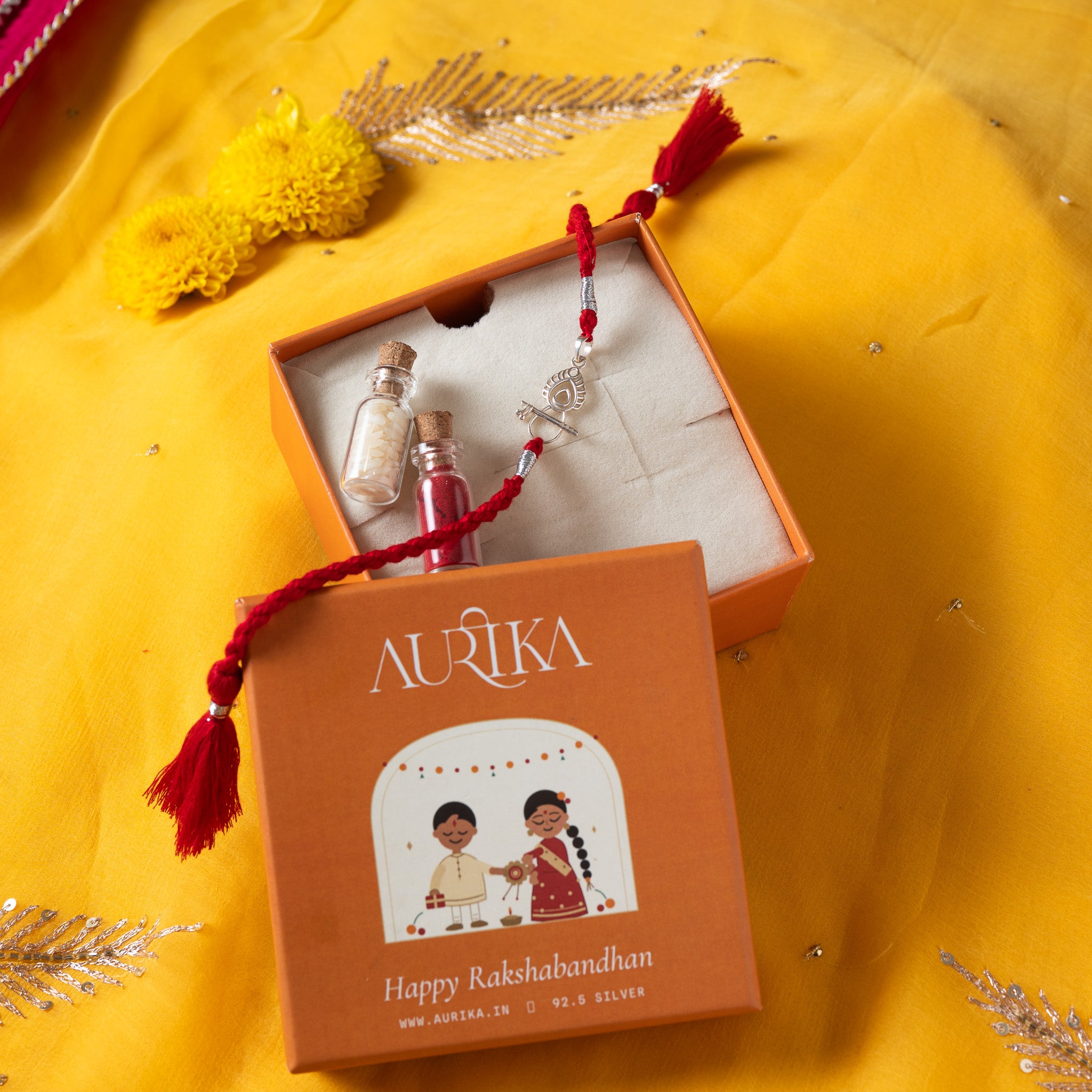 Bansuri and peacock feather rakhi with its marigold cord in an open Aurika gift box on rani pink cloth