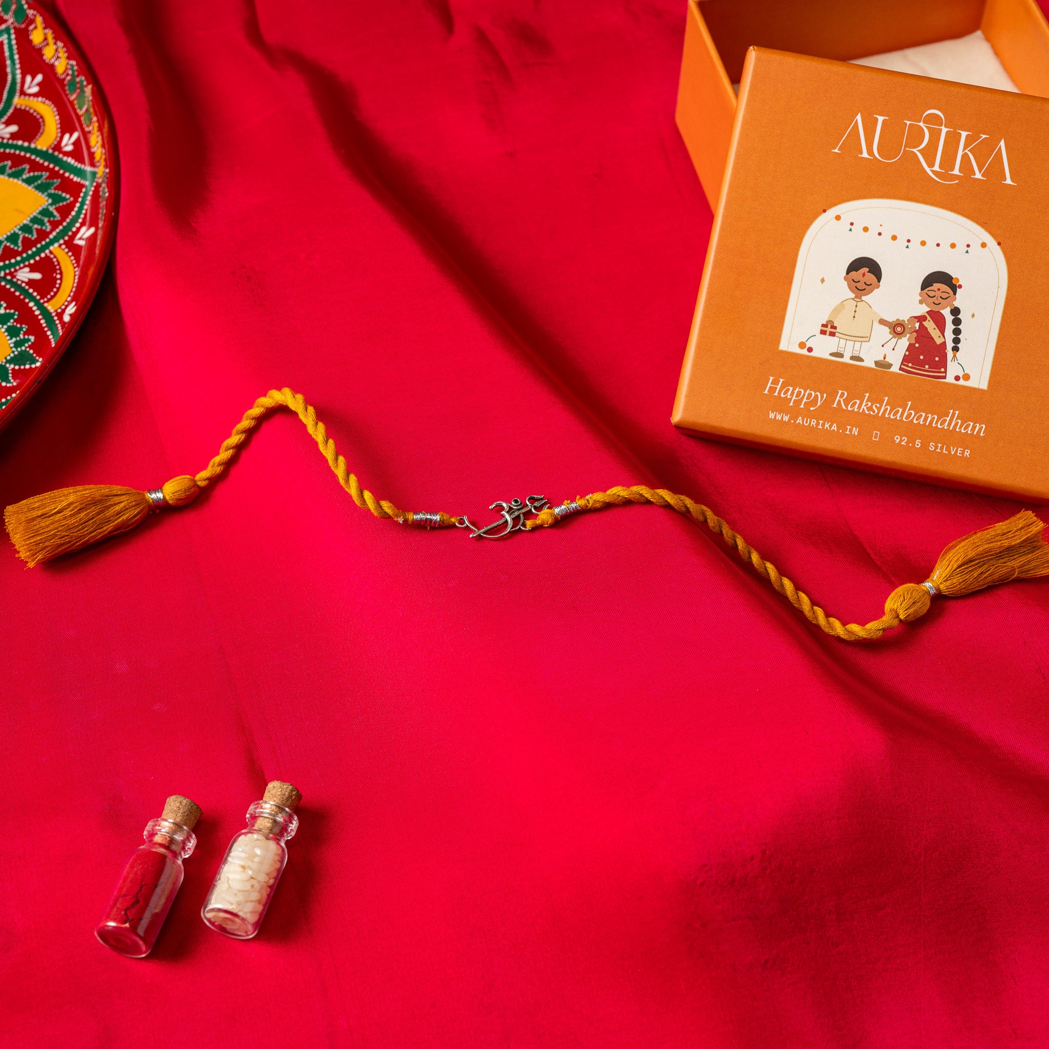 Om with a trishul rising from it in 925 silver on a red twisted silk rakhi cord with tassels