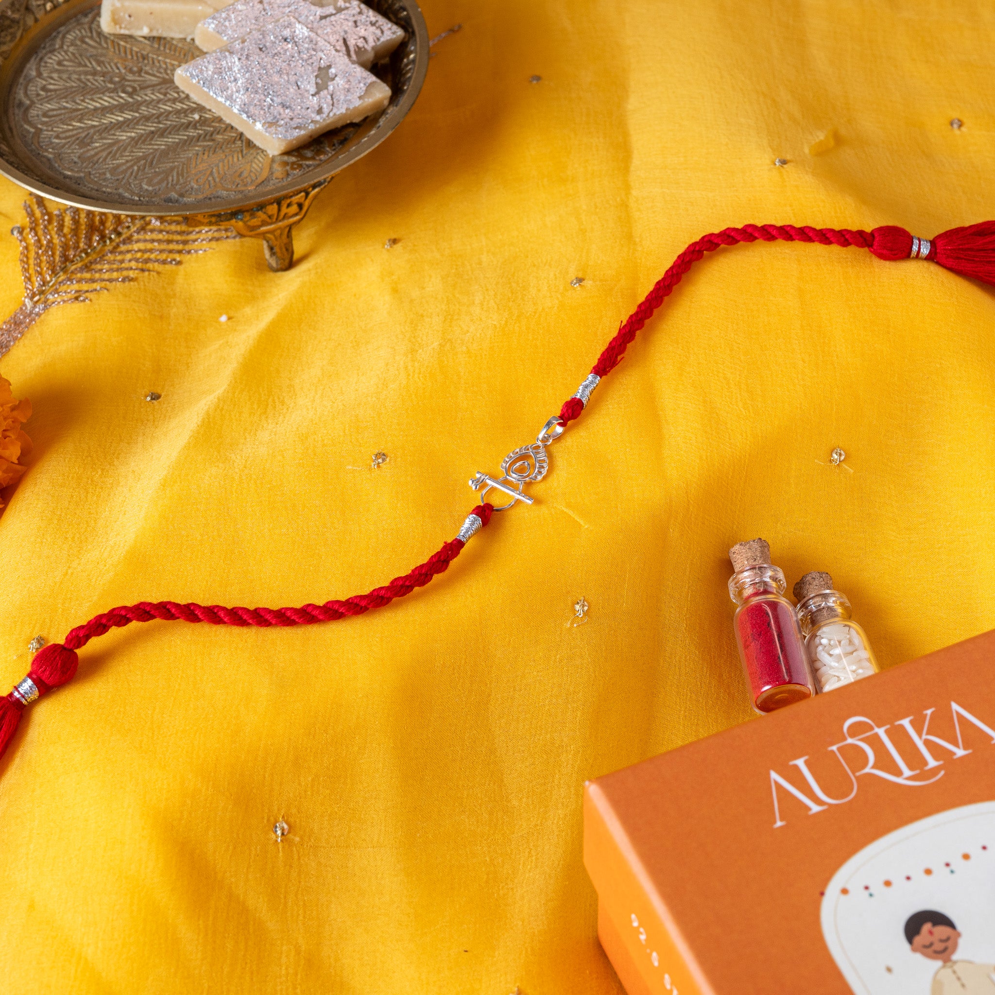 Bansuri laid across a peacock feather in 925 silver on a red twisted silk rakhi cord with tassels