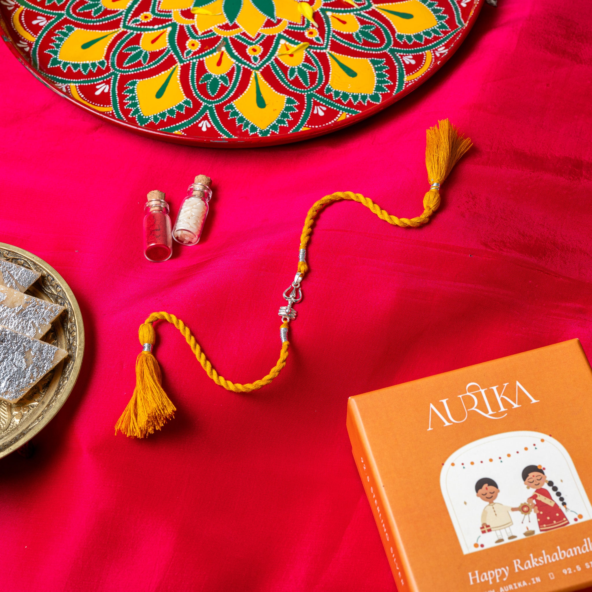 Trishul and shivling rakhi in 925 silver on a marigold yellow silk cord, on rani pink cloth