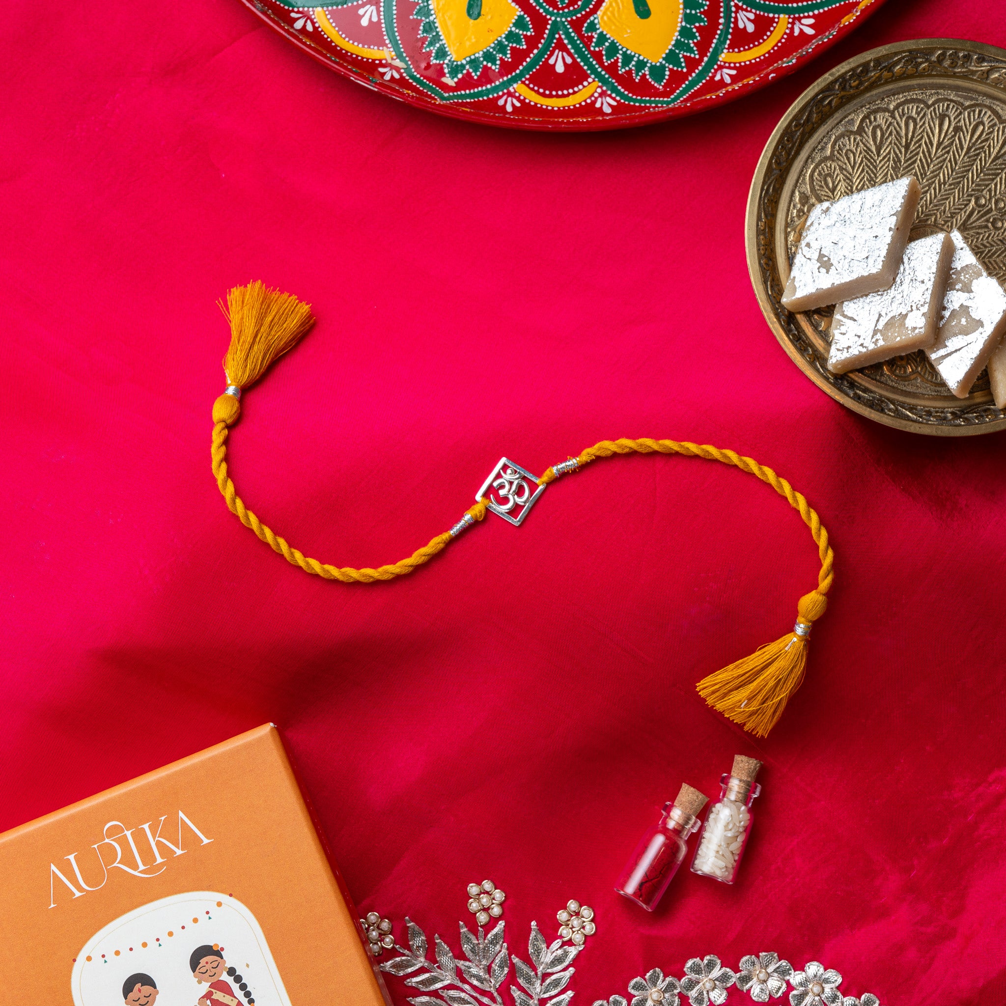 Om square rakhi in 925 silver on a marigold yellow silk cord, on rani pink cloth