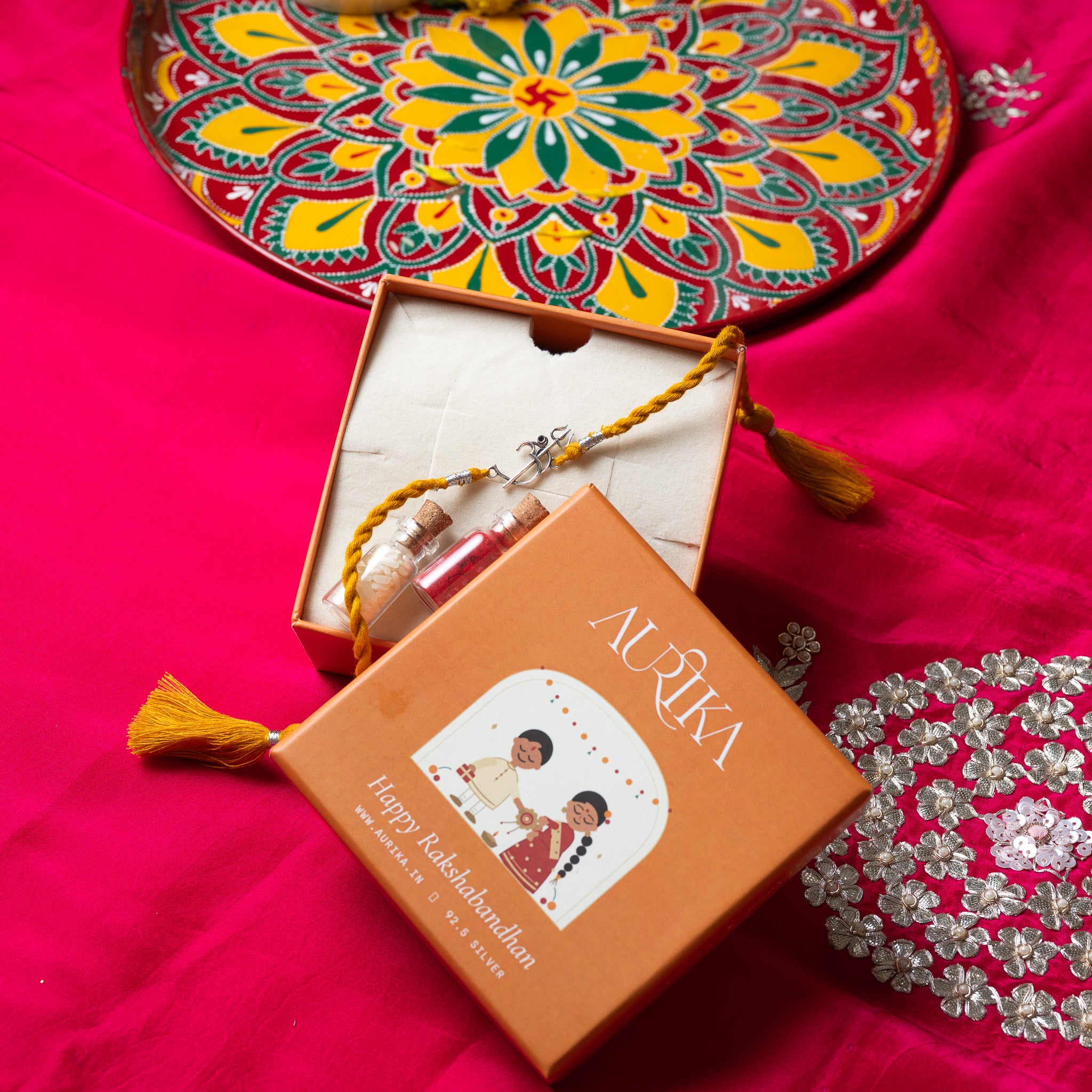 Om trishul rakhi with its marigold cord in an open Aurika gift box on rani pink cloth