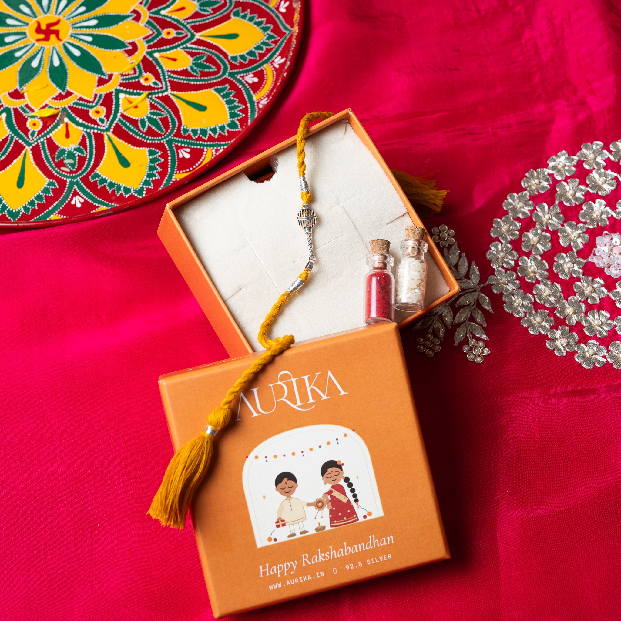 Gada mace head rakhi with its red cord in an open Aurika gift box with roli and akshat vials
