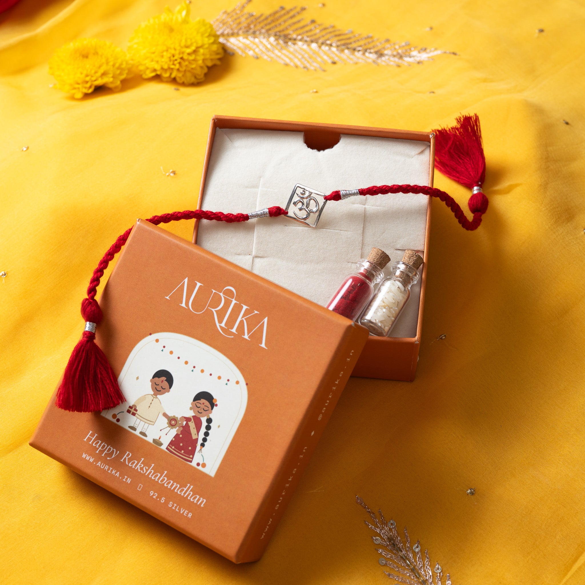 Om square rakhi with its red cord in an open Aurika gift box with roli and akshat vials