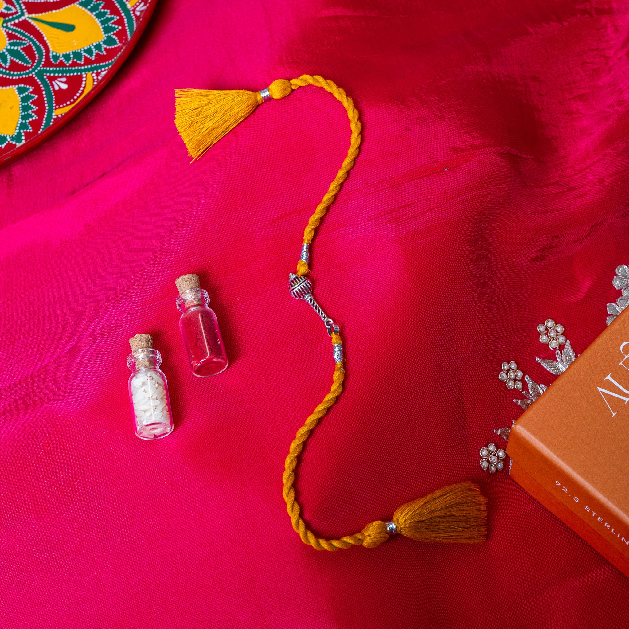 Gada mace head rakhi in 925 silver on a marigold yellow silk cord, on rani pink cloth