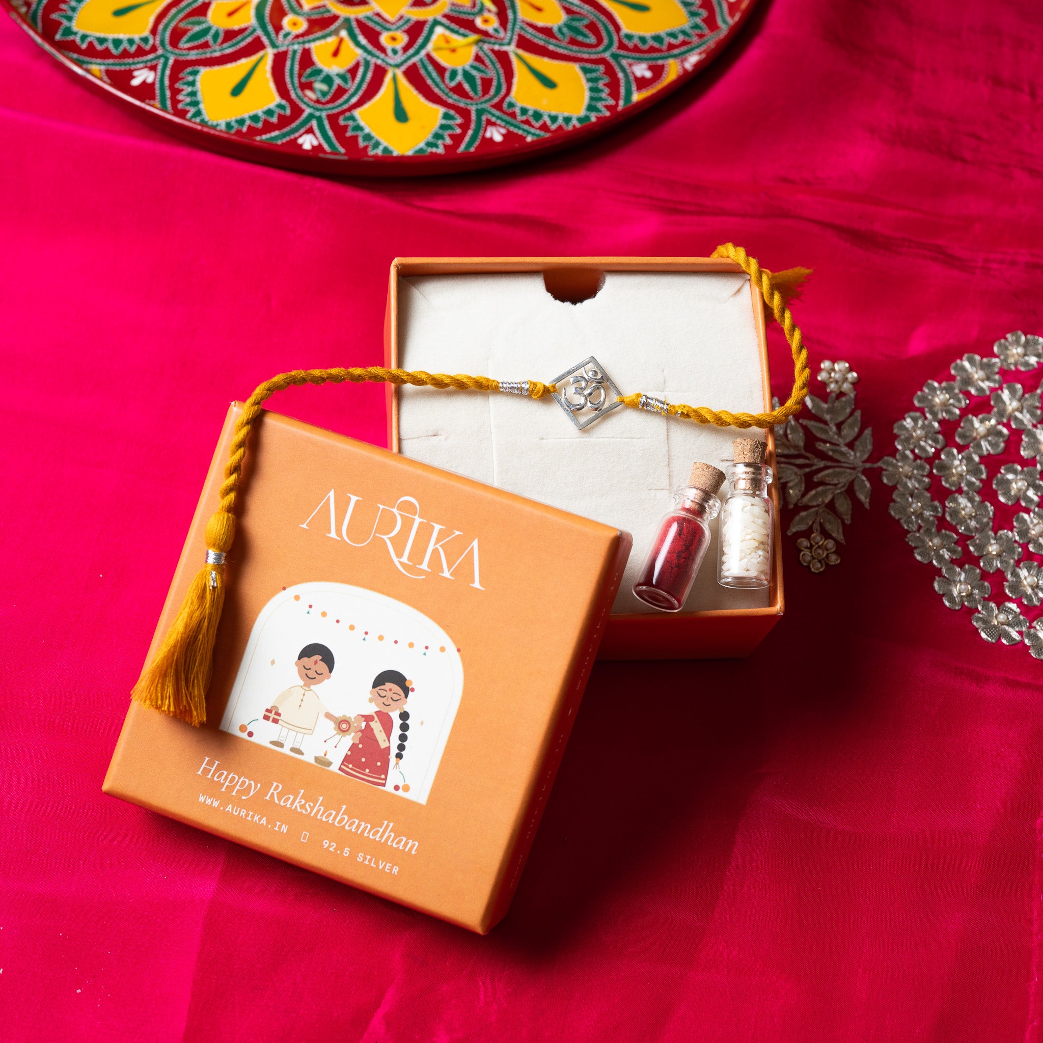 Om square rakhi with its marigold cord in an open Aurika gift box on rani pink cloth