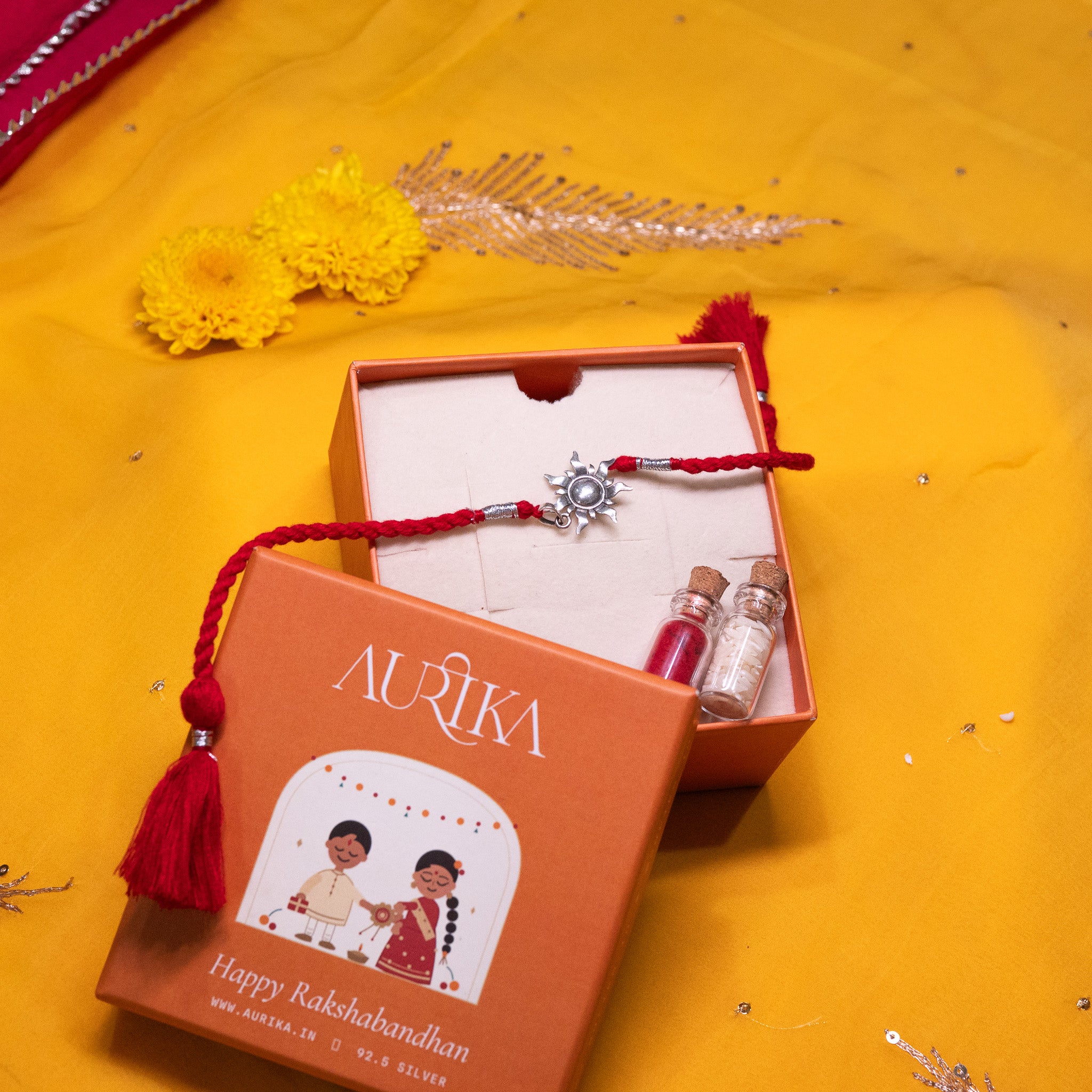 Surya sunburst rakhi with its red cord in an open Aurika gift box with roli and akshat vials