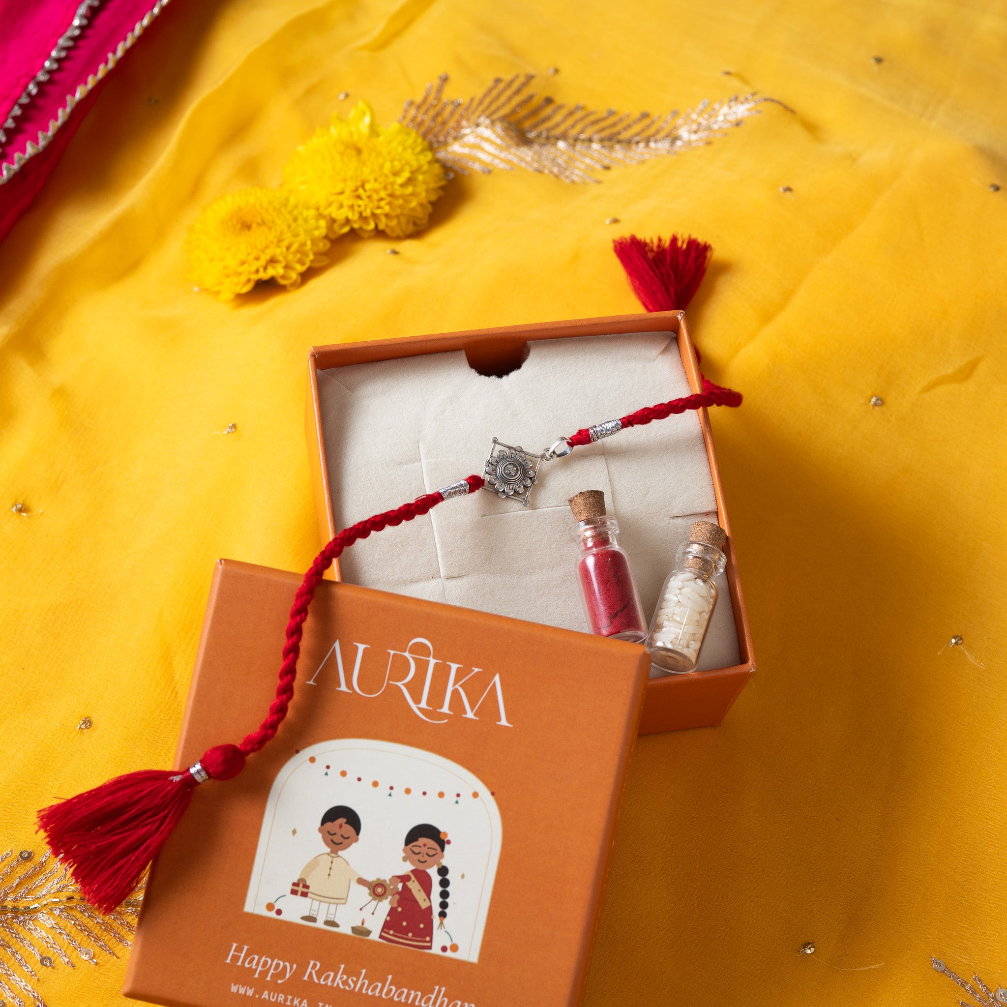 Champak flower rakhi with its marigold cord in an open Aurika gift box on rani pink cloth