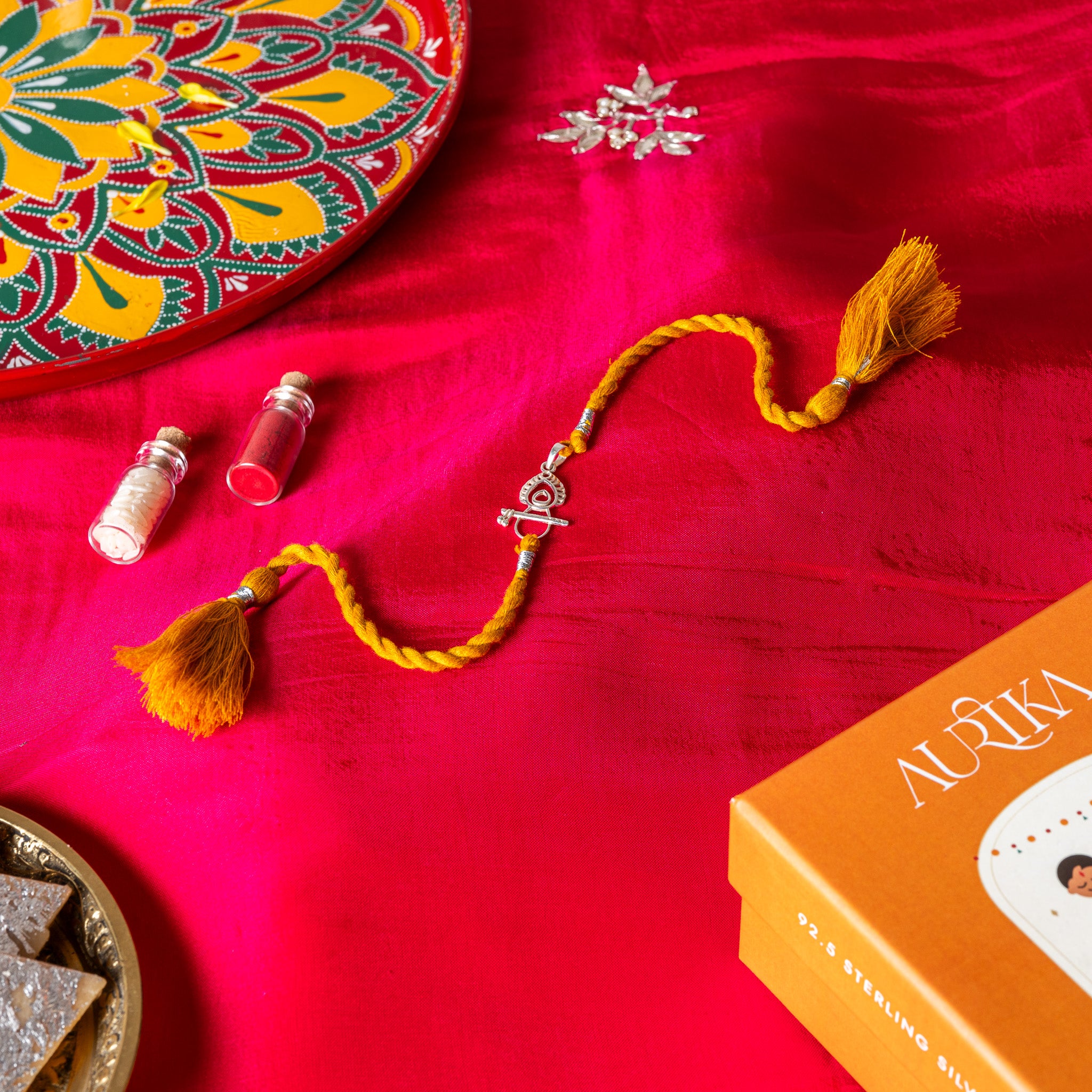 Bansuri and peacock feather rakhi in 925 silver on a marigold yellow silk cord, on rani pink cloth